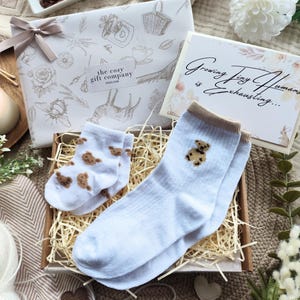 May include: A gift set featuring baby socks with teddy bear designs, a decorative box, and a card that reads "Growing Tiny Humans is Exhausting...". The set includes a wrapped gift with a bow, a lit candle, and decorative flowers.