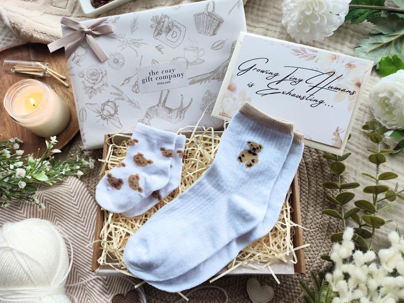 May include: A gift set featuring baby socks with teddy bear designs, a decorative box, and a card that reads "Growing Tiny Humans is Exhausting...". The set includes a wrapped gift with a bow, a lit candle, and decorative flowers.