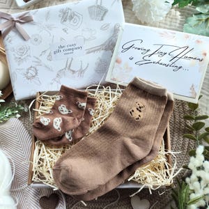 May include: A gift box with baby socks, a candle, and a card that reads "Growing Tiny Humans is Exhausting...". The socks are brown with teddy bear designs. The gift box is wrapped in white paper with a bow.