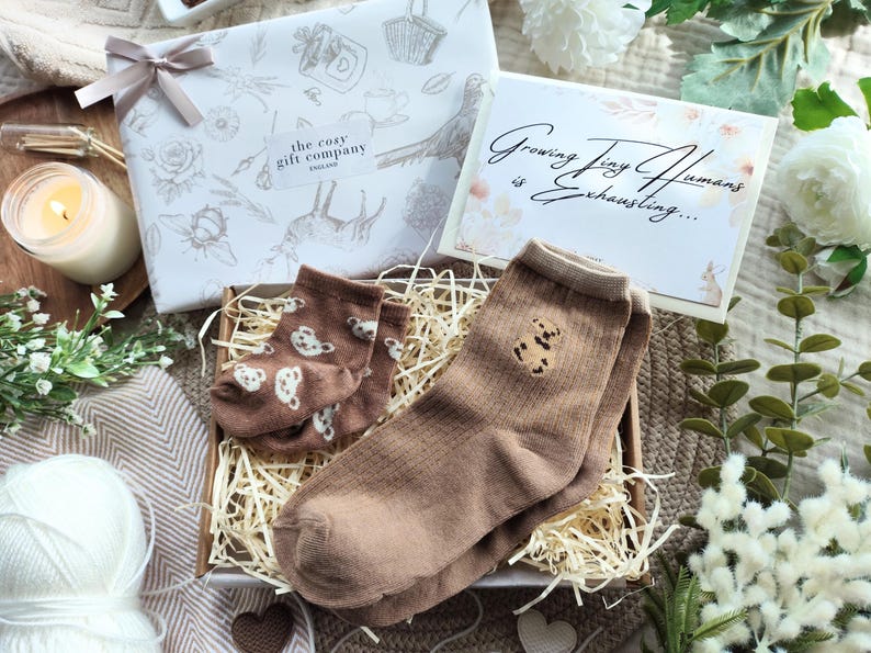 May include: A gift box with baby socks, a candle, and a card that reads "Growing Tiny Humans is Exhausting...". The socks are brown with teddy bear designs. The gift box is wrapped in white paper with a bow.