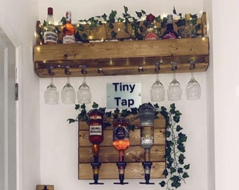 May include: A wooden bar cart with a shelf for bottles and glasses. The cart has a sign that says "Tiny Tap" and a bottle opener that says "Take your top off!" There are three bottles of liquor with spouts and a row of shot glasses on the counter below.
