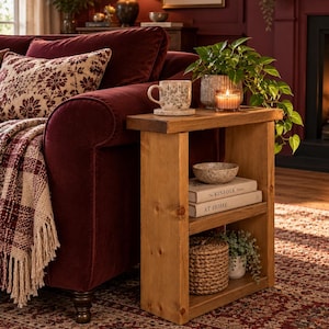 May include: A rectangular wooden side table with two shelves, featuring a natural wood grain finish. The top shelf holds a patterned mug, a potted plant, and a candle. The lower shelf contains a speckled bowl, two books titled The Kinfolk Home and At Home, a woven basket, and a small plant.