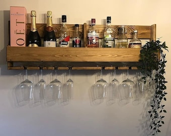 Könnte beinhalten: Ein wandmontierter Weinständer aus Holz mit einem Regal und einem Hängeregal für Weingläser. Der Weinständer ist mit Flaschen Wein und Spirituosen gefüllt, darunter eine Flasche Moët & Chandon Champagner. Eine kleine Topfpflanze hängt am Weinständer.