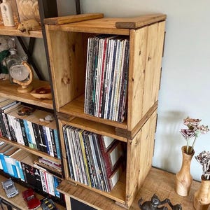 May include: Two stacked, light-brown wooden record storage cubes. Each cube is filled with vinyl records. The cubes are made of unfinished wood and have a rustic appearance. The cubes are sitting on a wooden surface.