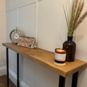 May include: A wooden console table with black metal legs. It features a clock, a weekly planner, a candle, and a vase with decorative grass. The table is against a white panelled wall.