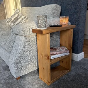 May include: A small, light brown wooden side table with a shelf, holding two books. A cup with the word "tea" printed on it sits on the table, along with a candle in a glass. The table is next to a grey sofa.