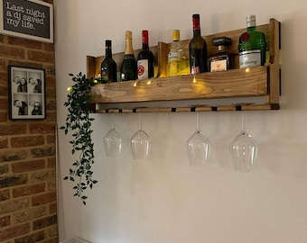 May include: A wooden wall-mounted wine rack with a shelf and glass holders. The rack is stained a dark brown colour and has a rustic look. There are six wine bottles and four wine glasses on the rack. The rack is mounted on a white wall with a brick wall to the left.