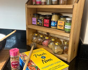 Puede incluir: Un especiero de madera lleno de varios frascos y botellas de especias. Un libro de cocina titulado "Pinch of Nom" está junto a una botella de condimento Batts Piri Piri. También se ve un molinillo de especias de madera.