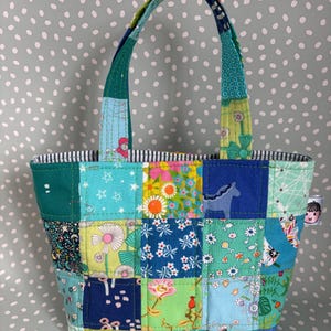 May include: A patchwork tote bag in shades of blue, green, and yellow. The bag features a variety of fabric squares, a striped lining, and matching handles. A small tag with a cartoon face is attached to the side.