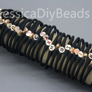 May include: A stack of black elastic bracelets with gold and white letter beads. Each bracelet has a single letter bead with a colorful enamel design.