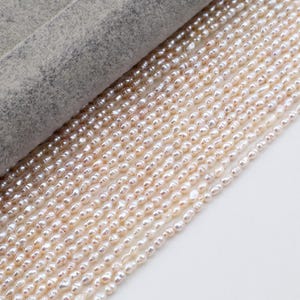 Puede incluir: Primer plano de múltiples hilos de perlas de agua dulce ovaladas en tonos blanco y marrón claro. Las perlas están ensartadas, creando una apariencia delicada. Las perlas se muestran sobre una superficie blanca.