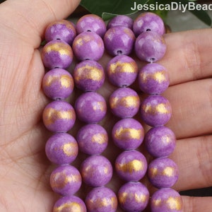 May include: A strand of purple and gold round beads. The beads are made of a smooth, polished material and have a subtle shimmer. The beads are strung on a clear elastic cord.
