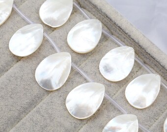May include: Close-up of a strand of pearly white teardrop-shaped beads. The beads have a lustrous, iridescent sheen and are strung on a clear, thin cord. The beads are arranged on a gray, textured surface.