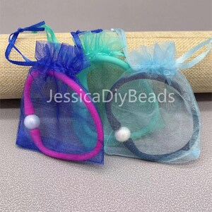 May include: Three small organza bags with drawstrings, each containing a hair tie and a pearl bead. The bags are blue, teal, and pink.