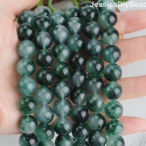 May include: Strand of dark green, round beads. The beads have a polished, translucent appearance with variations in color, ranging from deep emerald to nearly black. The beads are held in a hand, showcasing their size and texture. The beads are likely for jewelry making.