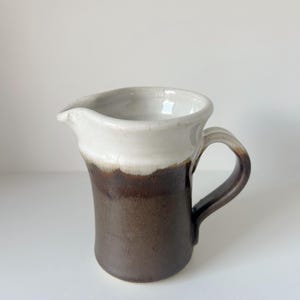 Può includere: Brocca in ceramica fatta a mano con bordo e manico bianchi, che sfuma in un corpo marrone scuro. La brocca ha un beccuccio per versare e un manico curvo. L'interno è bianco. Misure in centimetri.