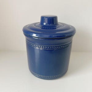 Könnte beinhalten: Ein kobaltblaues Keramikgefäß mit Deckel. Das Gefäß hat eine zylindrische Form mit einem dekorativen Band aus kleinen, erhabenen Punkten um die Mitte und den Boden. Der Deckel hat einen kleinen Griff und ein ähnliches Punktmuster. Das Gefäß steht auf einer weißen Oberfläche.