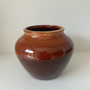 May include: A glazed ceramic vase in shades of brown. The vase has a wide, open mouth and a rounded body. The top rim is a lighter brown, transitioning to a darker brown at the base. The vase is likely for decorative use.
