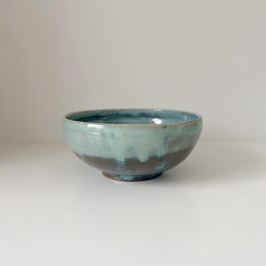 Könnte beinhalten: Eine handgefertigte Keramikschale mit glänzendem Finish. Die Schale zeigt einen Farbverlauf, der von einem dunkelbraunen Sockel zu einem hellen türkisblauen und blauen Rand übergeht. Die runde Schale hat ein schlichtes, elegantes Design, das sich zum Servieren von Speisen oder als Dekorationsstück eignet.