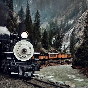 May include: A black steam locomotive train with a white number "18" on the side, pulling a string of orange passenger cars, travels through a mountain valley with a waterfall and a river.