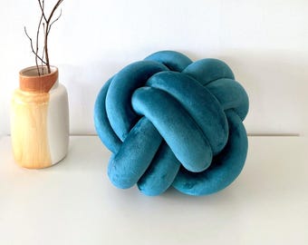 May include: A teal blue knot-shaped decorative pillow made of soft fabric. The pillow is on a white surface, next to a vase with a brown and yellow design and a branch with thin twigs.