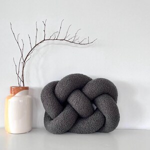 May include: A dark grey knotted cushion with a soft, textured surface. The cushion is shaped like a rectangle with rounded corners.