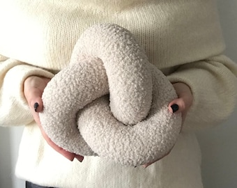 May include: A large, off-white, fluffy, knotted pillow. The pillow is tied in a knot shape and has a soft, fuzzy texture.