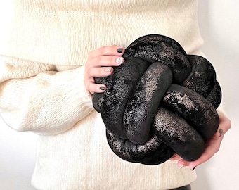 May include: A black velvet knot pillow with gold glitter accents. The pillow is being held by a person wearing a white sweater and black pants.