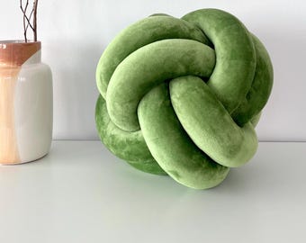 May include: A green velvet knotted pillow. The pillow is round and has a soft, plush texture. It is sitting on a white surface next to a vase with a brown and white design.