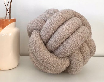 May include: A light brown, fluffy, knotted pillow. The pillow is round and has a soft, textured surface. It is sitting on a white surface next to a vase with a brown and white design.