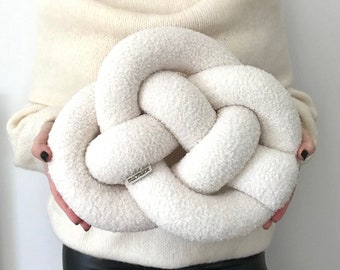May include: A large, white, fluffy, knotted pillow. The pillow is made of a soft, plush fabric and has a simple, modern design. The pillow has a small, silver tag with the word "macrame" on it.