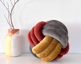 May include: A decorative knotted pillow with a gray, red, and yellow velvet fabric. The pillow is sitting on a white surface.