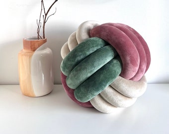 May include: A decorative pillow in the shape of a knot, with three colors: a deep green, a light beige, and a dusty rose. The pillow is sitting on a white surface.