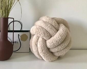 May include: A beige fluffy knotted pillow on a white surface. The pillow is made of soft, plush fabric and has a unique knotted design.