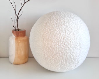 May include: A large white round pouf or cushion with a textured surface. The pouf is sitting on a white surface next to a vase with a brown and white design.
