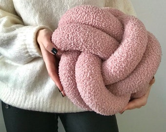 May include: A large, pink, fluffy knotted pillow. The pillow is made of a soft, fuzzy material and is shaped like a knot.