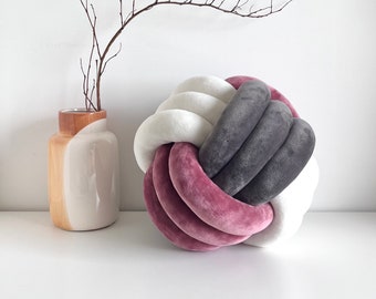 May include: A decorative pillow in the shape of a knot, made of three different colors of fabric: pink, gray, and white. The pillow is sitting on a white surface.