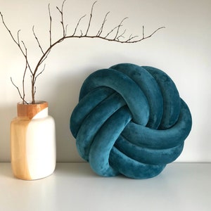 May include: A teal blue knotted pillow with a soft, plush texture. The pillow is round and has a decorative knot design.