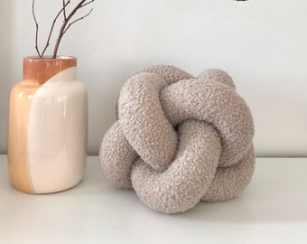 May include: A light brown, fluffy, knotted pillow on a white surface. A vase with a brown and white design holds a dried branch with twigs.