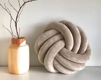 May include: A large, round, beige knotted pillow. The pillow is made of a soft, plush fabric and has a simple, modern design.