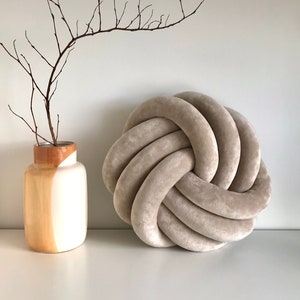 May include: A large, round, beige knotted cushion with a soft, plush texture. The cushion is made of a thick, woven fabric and has a simple, modern design.