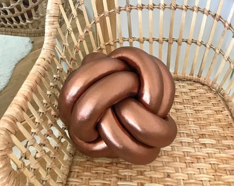 May include: A copper-colored knot pillow sits on a woven wicker chair. The pillow is made of a soft, plush fabric and has a unique knot design.