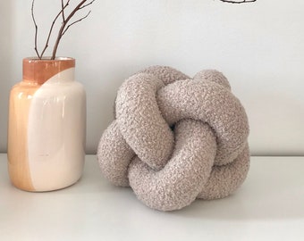 May include: A light brown, fluffy, knotted pillow. The pillow is sitting on a white surface. A vase with a brown and white design is in the background.