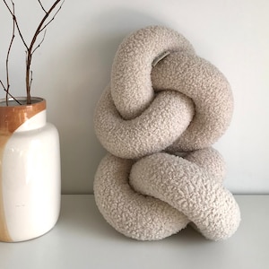 May include: A large, cream-colored, fluffy knotted pillow. The pillow is made of a soft, fuzzy material and is shaped like a knot. The pillow is sitting on a white surface.