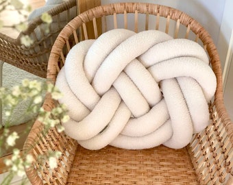 May include: A white knotted pillow sits on a woven wicker chair. The pillow is made of a soft, fluffy material and has a unique knotted design.