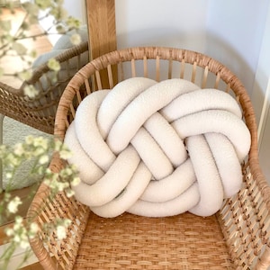 Può includere: Un cuscino bianco annodato si trova su una sedia in vimini intrecciato. Il cuscino è realizzato in un materiale morbido e soffice e presenta un design annodato unico.