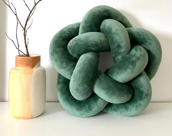 May include: A green velvet knotted pillow in the shape of a star. The pillow is sitting on a white surface next to a vase with a brown and white design.