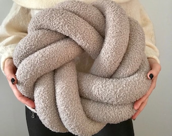 May include: A large, knotted pillow made of soft, light brown fabric. The pillow is shaped like a large knot and has a fluffy, textured surface.