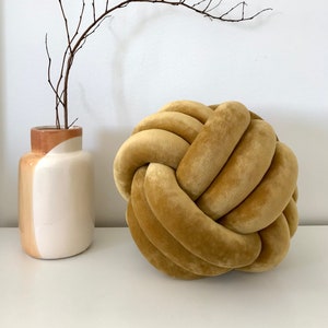 Puede incluir: Un cojín grande, redondo, de terciopelo amarillo mostaza con nudos. El cojín está hecho de múltiples hebras de tela que están anudadas para crear un diseño único y decorativo.