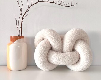May include: A white fluffy knotted pillow on a white surface. A brown twig branch in a white and orange vase is in the background.